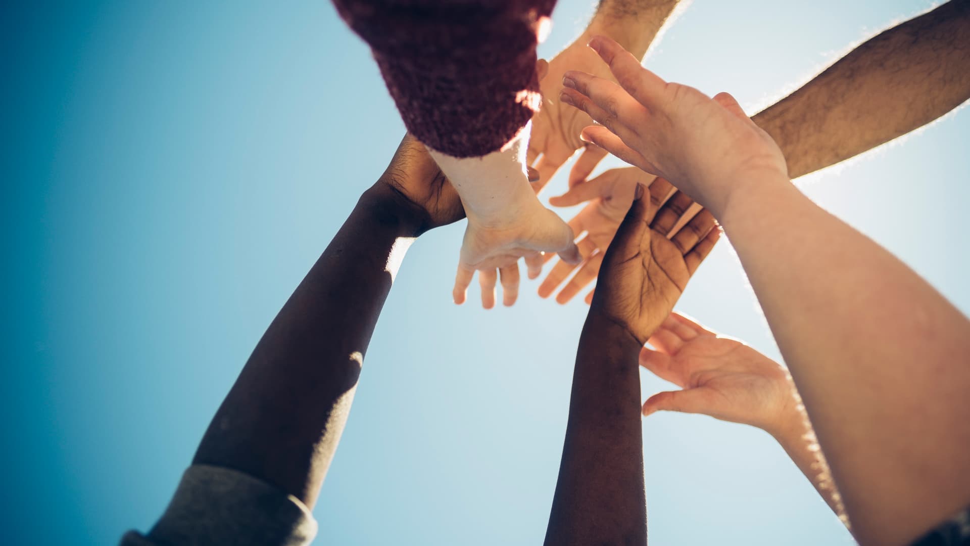 En gruppe ulike hender som strekker seg opp og slår seg sammen mot en knallblå himmel, som symboliserer samhold, teamarbeid og samarbeid.