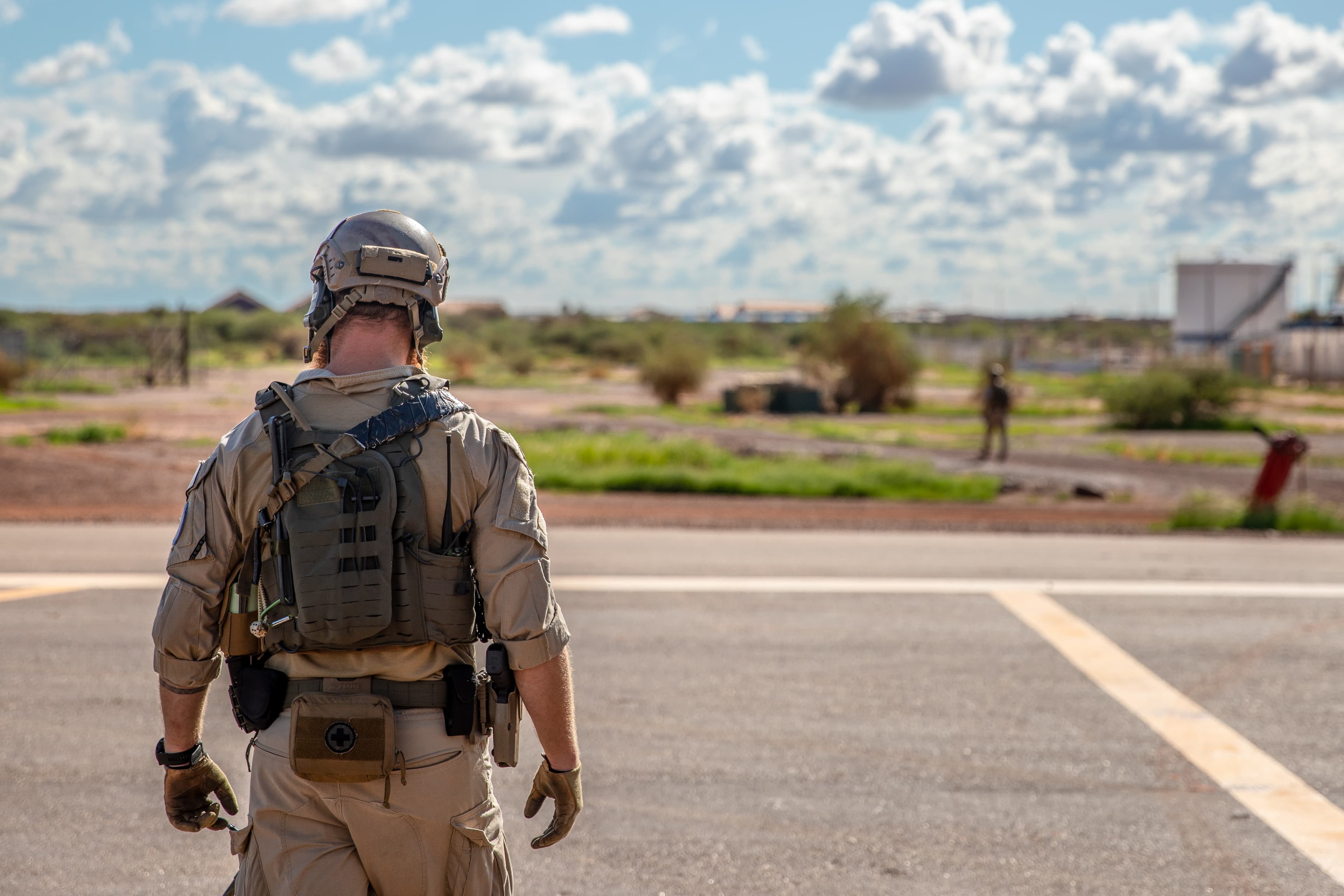 En soldat i taktisk utstyr står på en flyplassbane, med ryggen til kameraet, med åpent landskap, en annen soldat og en overskyet himmel i bakgrunnen.