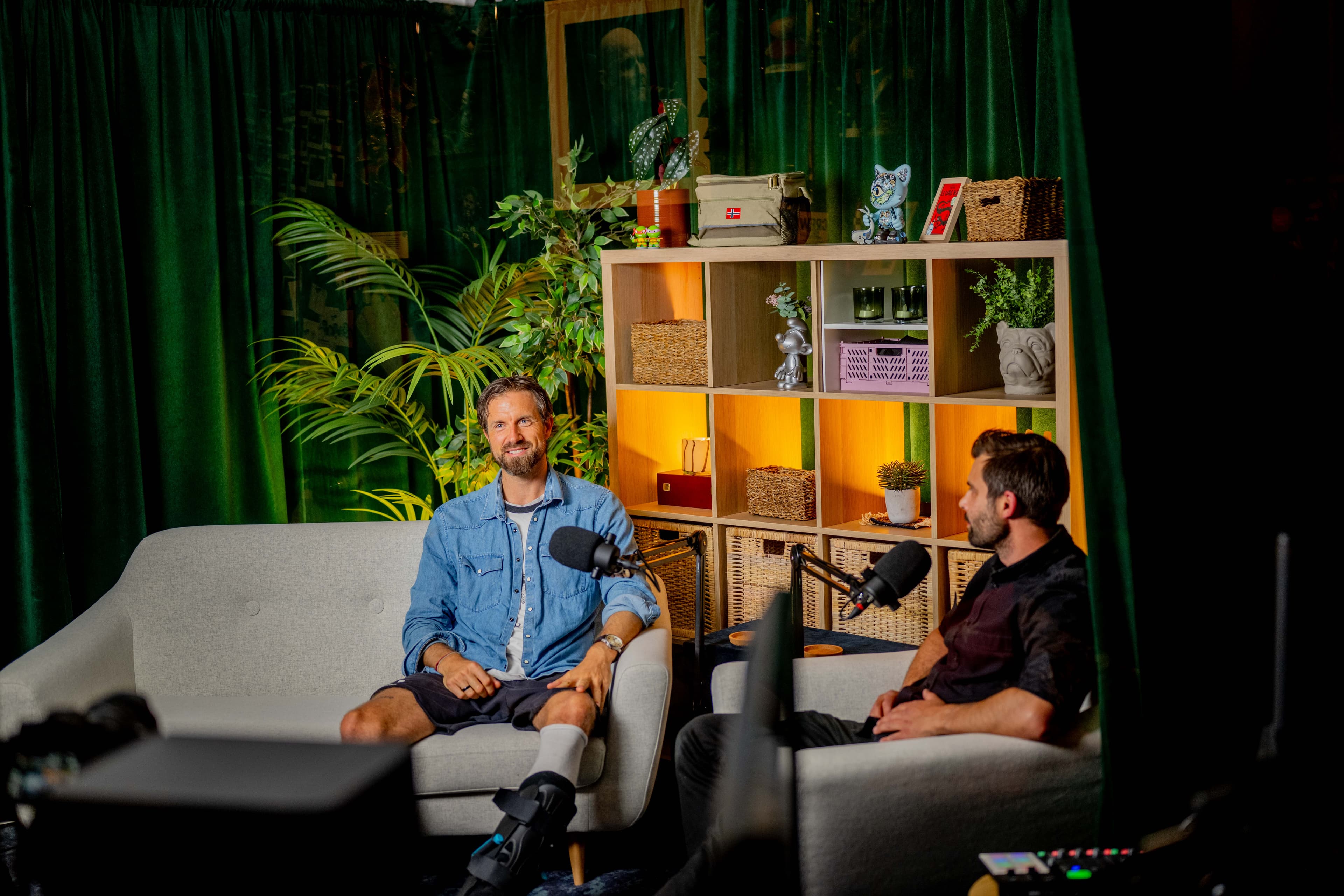 To menn sitter overfor hverandre i en sofa og en stol og snakker i mikrofoner i et koselig, plantefylt podcaststudio med grønne gardiner og en trehylle med bøker, planter og dekor bak dem.