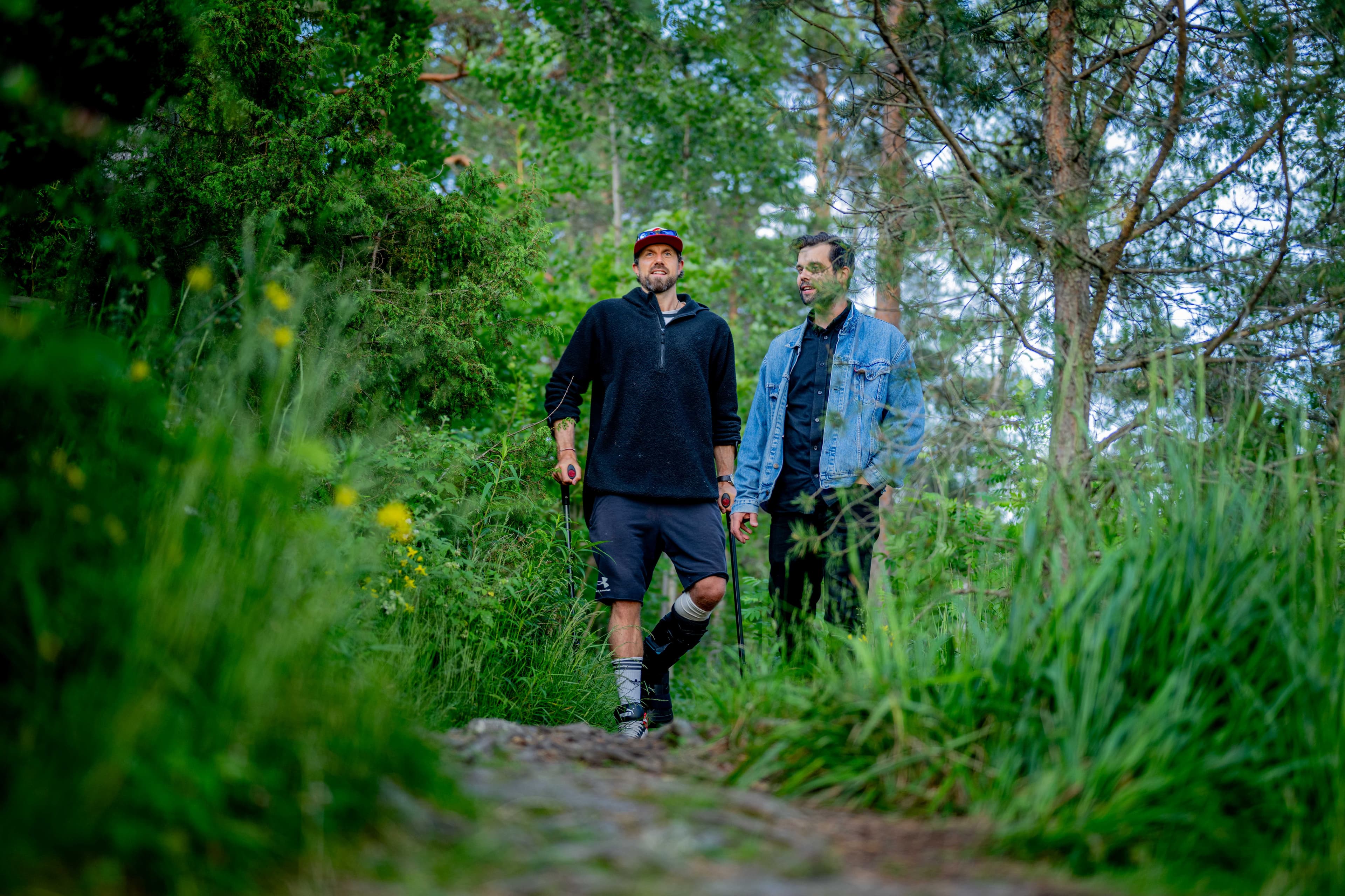 To menn går sammen på en skogssti omgitt av frodige, grønne trær og planter. Den ene har på seg caps, svart genser og shorts, mens den andre har briller og dongerijakke. Scenen er fredfull og full av natur.