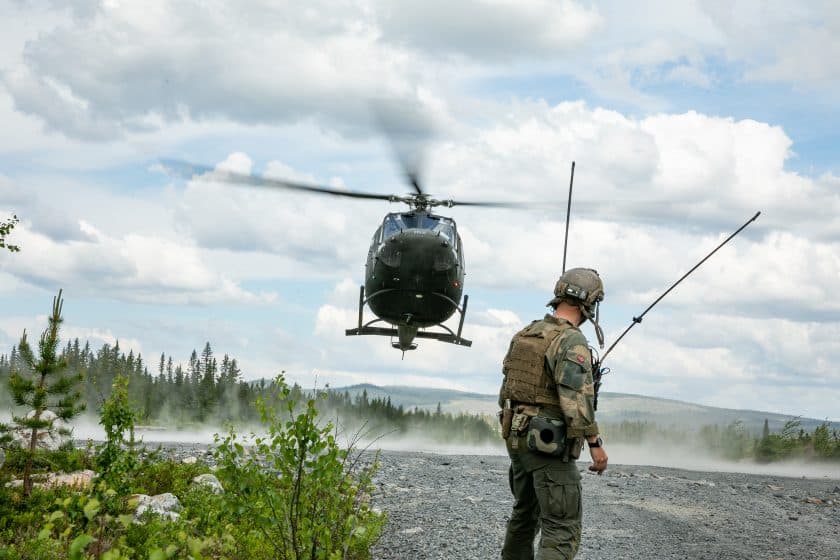 En soldat i uniform og utstyr står på en steinete bakke med et helikopter svevende i nærheten, omgitt av trær og en skyet himmel.