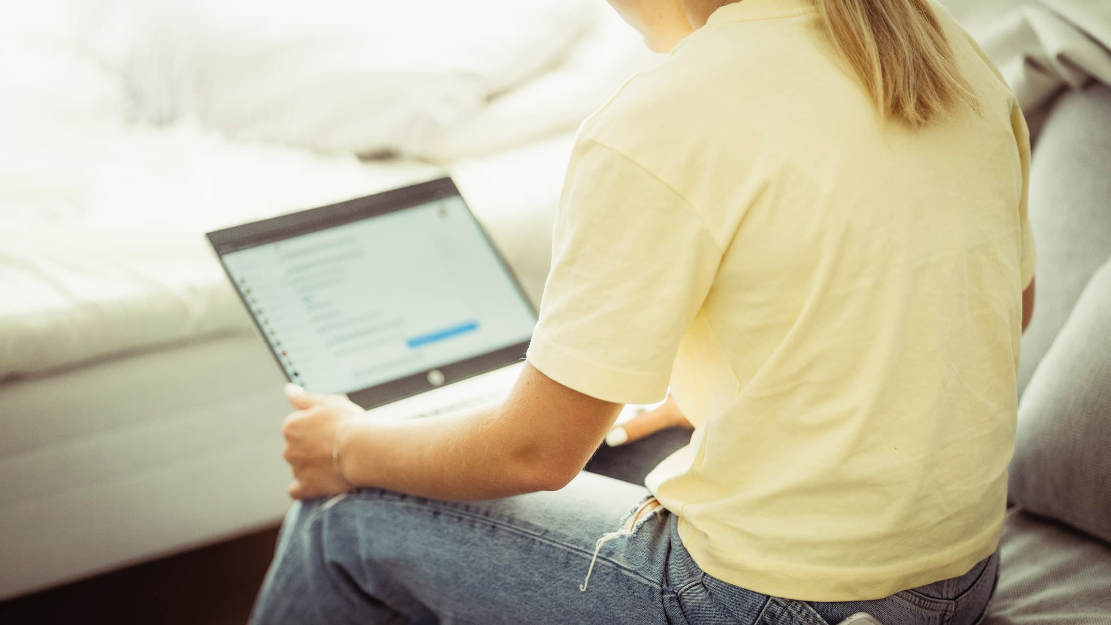 En person med langt blondt hår, iført gul skjorte og blå jeans, sitter på en seng med en åpen bærbar PC, og en uskarp dataskjerm viser en liste eller meldinger.