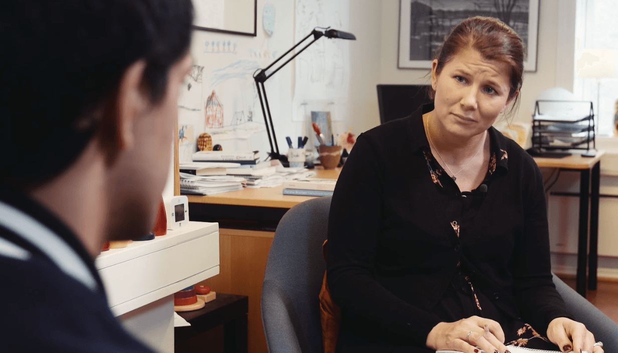 En kvinne sitter på et kontor og lytter oppmerksomt og tar notater under en samtale med en person som står med ryggen til kameraet. Kontoret har et skrivebord, en lampe, papirer og kunstverk på veggene.