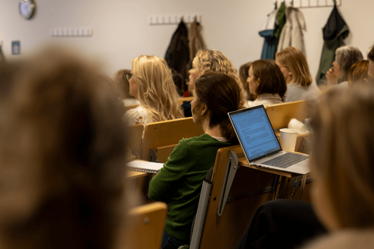 En gruppe mennesker sitter i et klasserom, vendt mot fronten. En person har en bærbar PC åpen på pulten, med et dokument på skjermen. Jakker henger på knagger i bakgrunnen. Stemningen virker konsentrert og oppmerksom.