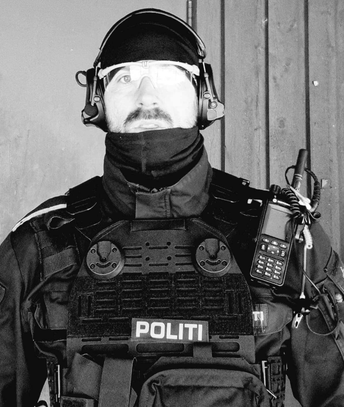 En person iført taktisk utstyr og hodetelefoner, med en radio festet til vesten merket POLITI, står foran en trevegg. Personen bærer også vernebriller og ansiktsdekkende plagg.