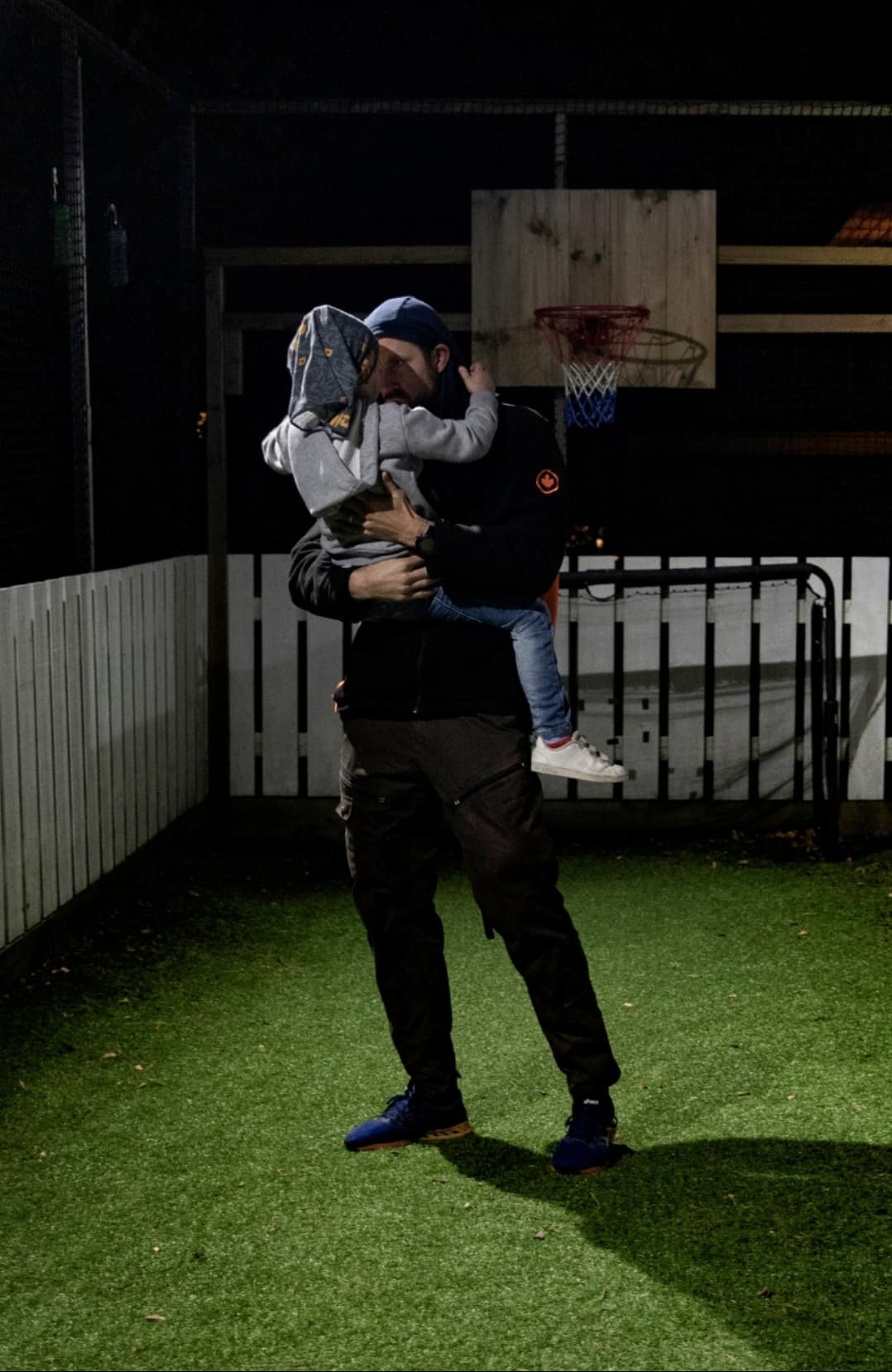 En person står på kunstgresset om natten og holder et lite barn i hettegenser og jeans. Bak dem er det en basketballkurv og et hvitt gjerde. Scenen er svakt opplyst.