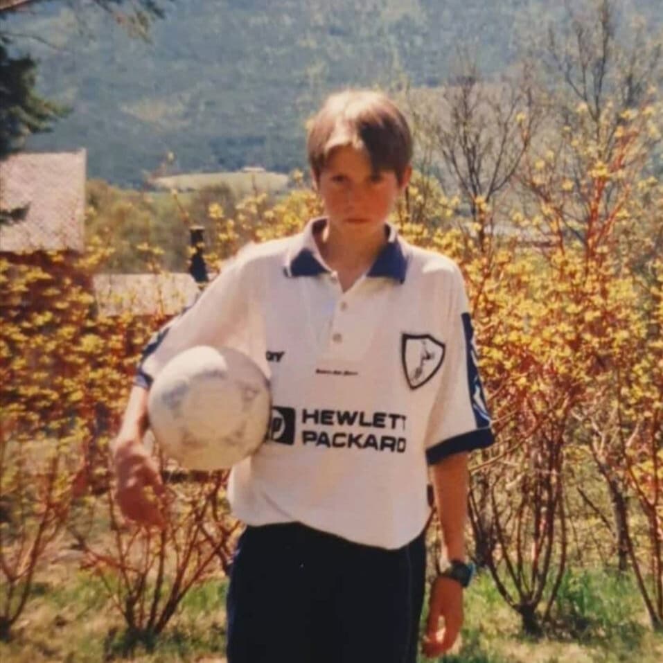 En ung gutt står utendørs med en fotball i hånden, iført en fotballtrøye med Hewlett Packard på forsiden. Bak ham er det busker med gule blomster og utsikt til fjell og trær i bakgrunnen.
