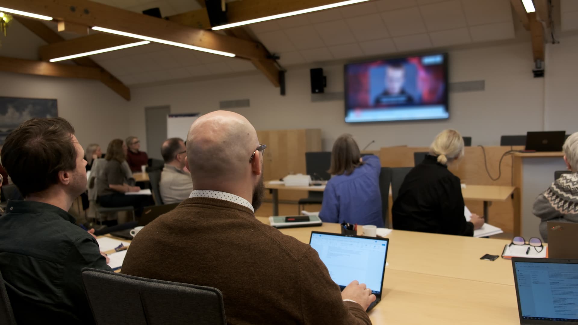 Folk sitter i et konferanserom, noen med bærbare datamaskiner, mens de ser på en uskarp nyhetssending på en stor skjerm foran i rommet. Settingen virker profesjonell og fokusert.