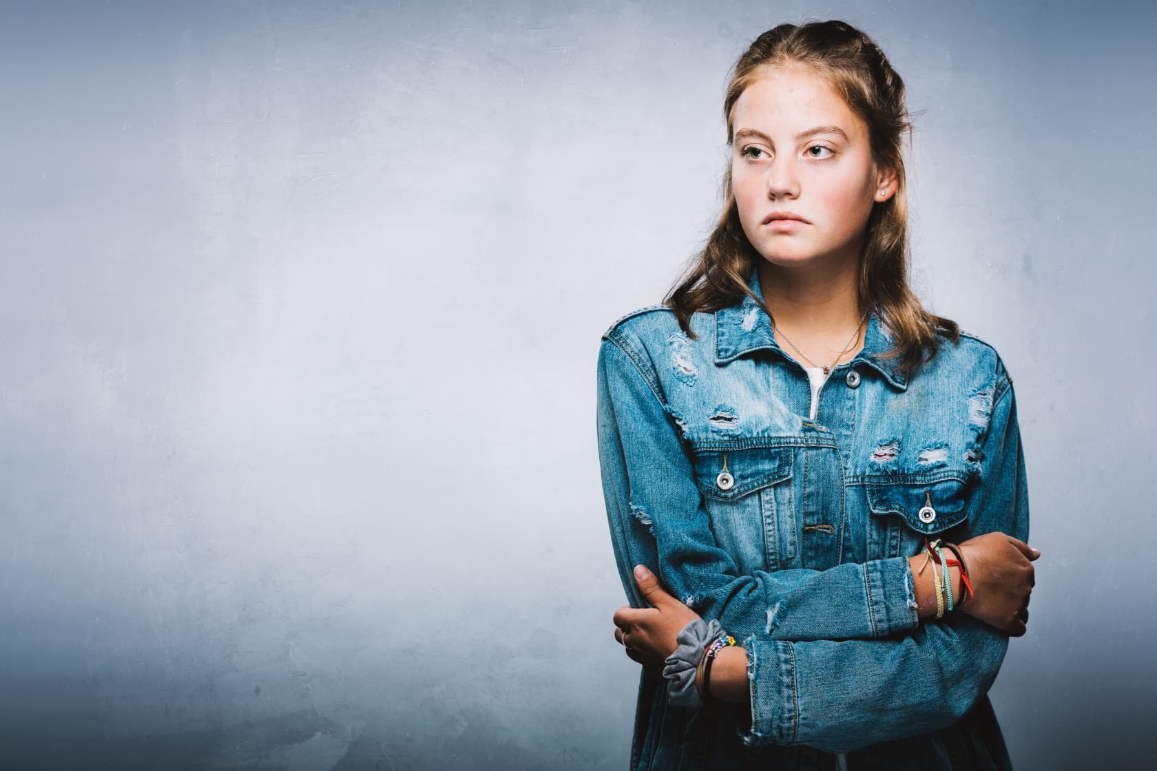 Tenåringsjente med langt brunt hår og en slitt dongerijakke står med armene i kors og ser til siden med et alvorlig uttrykk, mot en ensfarget lyseblå bakgrunn.