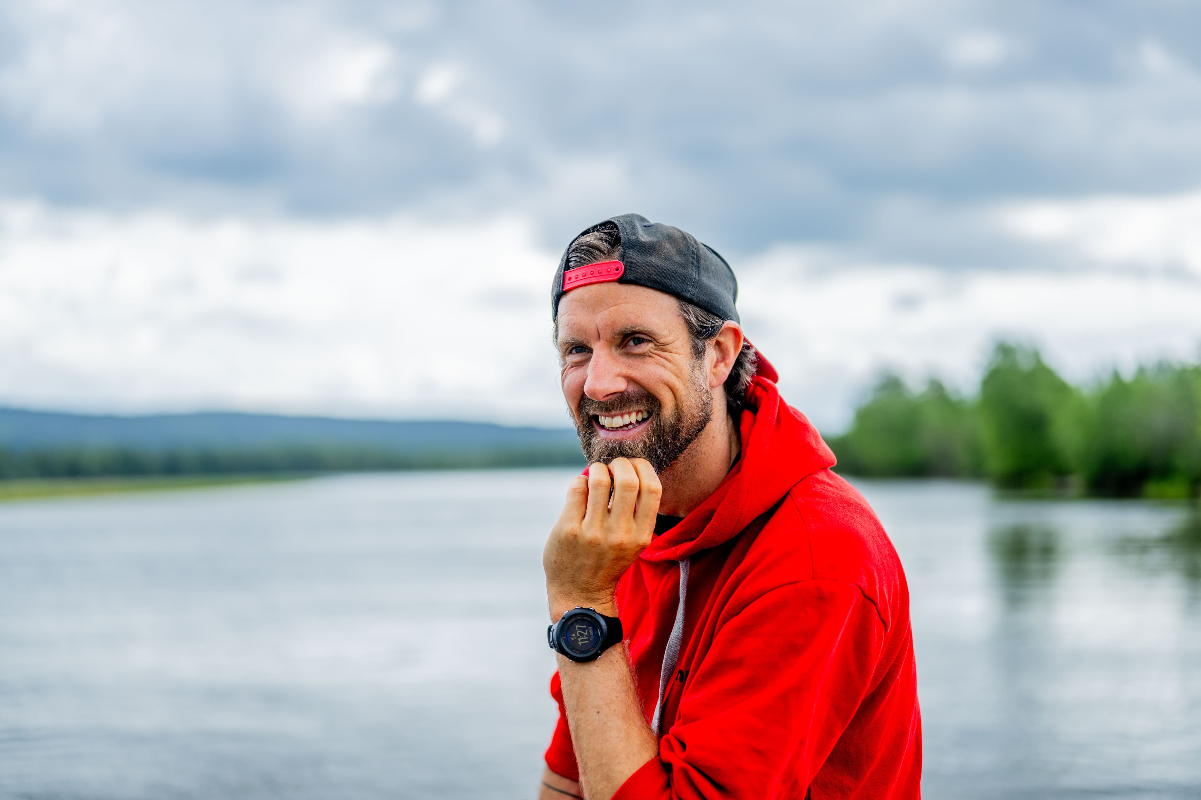 En mann i rød hettegenser og baklengs caps smiler mens han står ved en rolig innsjø, med trær og åser i bakgrunnen under en overskyet himmel.