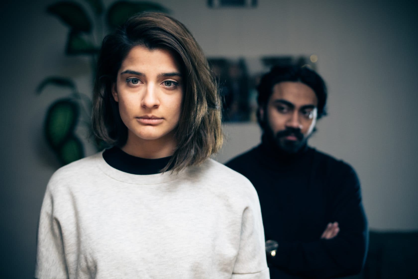 En kvinne står i forgrunnen med et alvorlig uttrykk, mens en mann i bakgrunnen ser på henne med armene i kors, noe som tyder på spenning eller konflikt mellom dem.