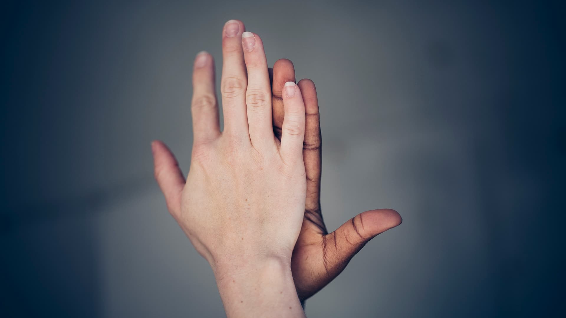 Nærbilde av to hender, en lys og en mørk, presset håndflate mot håndflate mot en nøytral, uskarp bakgrunn.
